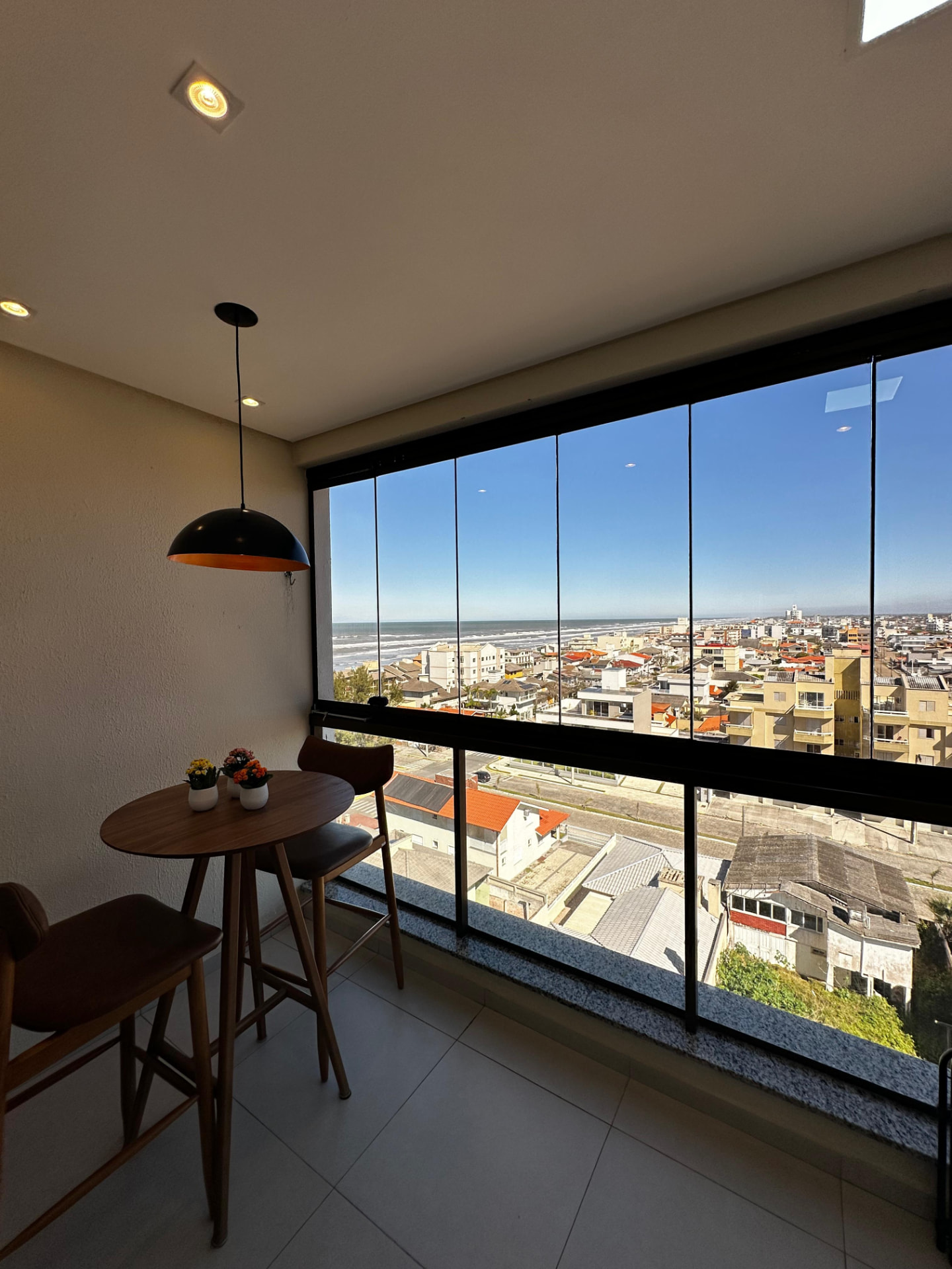 Sacada fechada com vidro, mesa alta e vista para o mar do Balneário Arroio do Silva