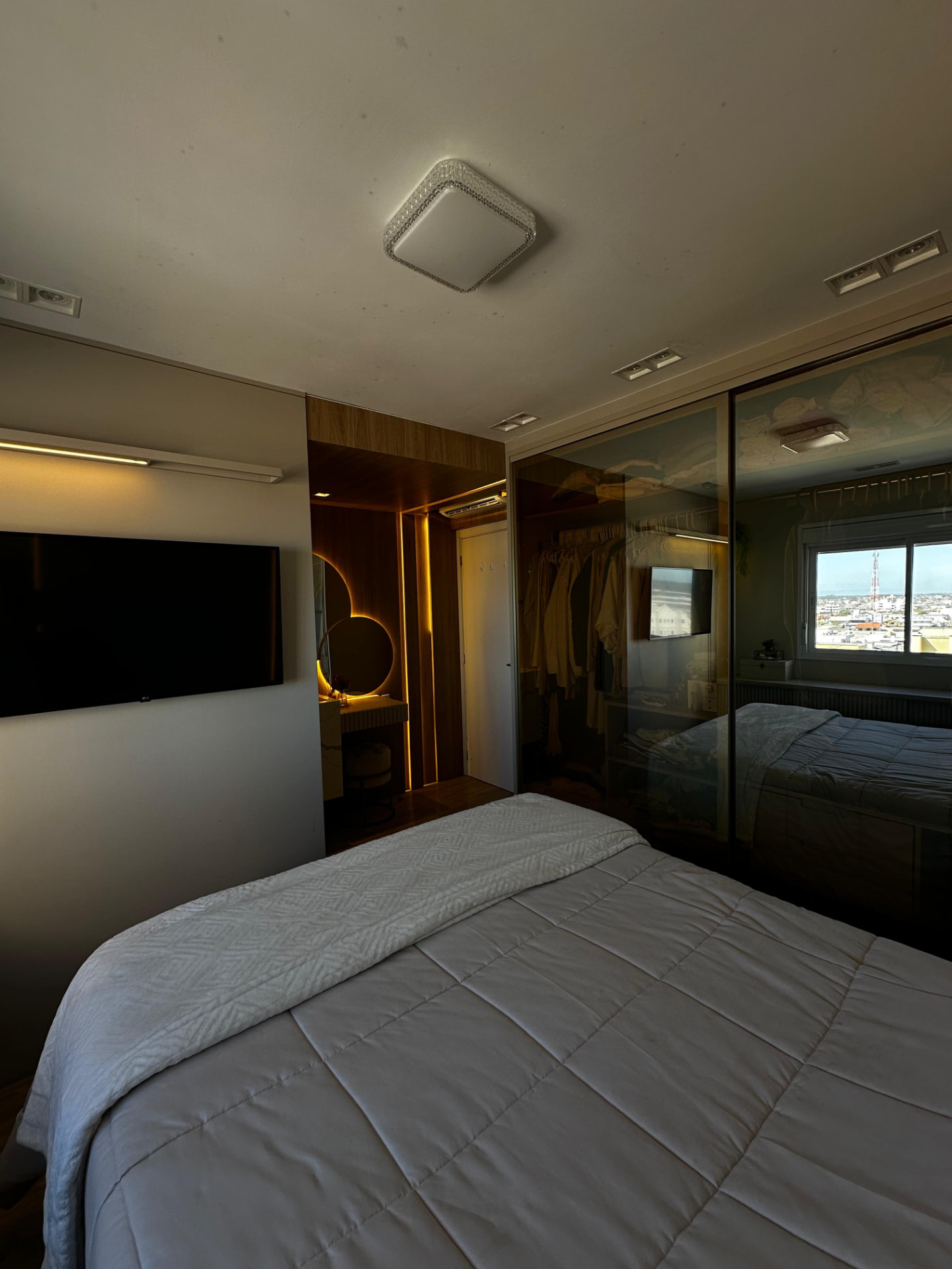Suíte com TV, closet com portas de vidro bronze e vista para a cidade